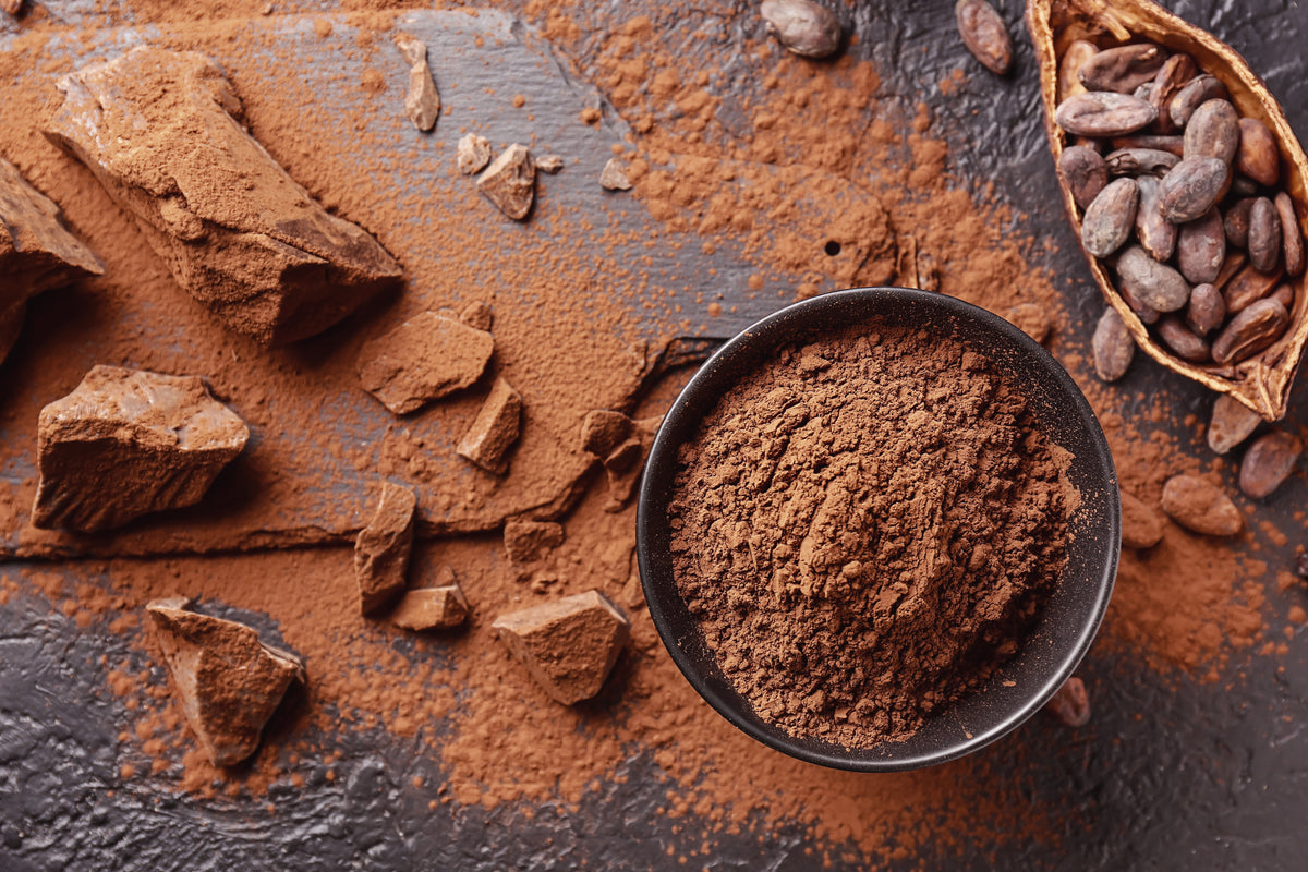 Poudre de cacao dans un bol noir, éclats de cacao et cabosse ouverte sur ardoise