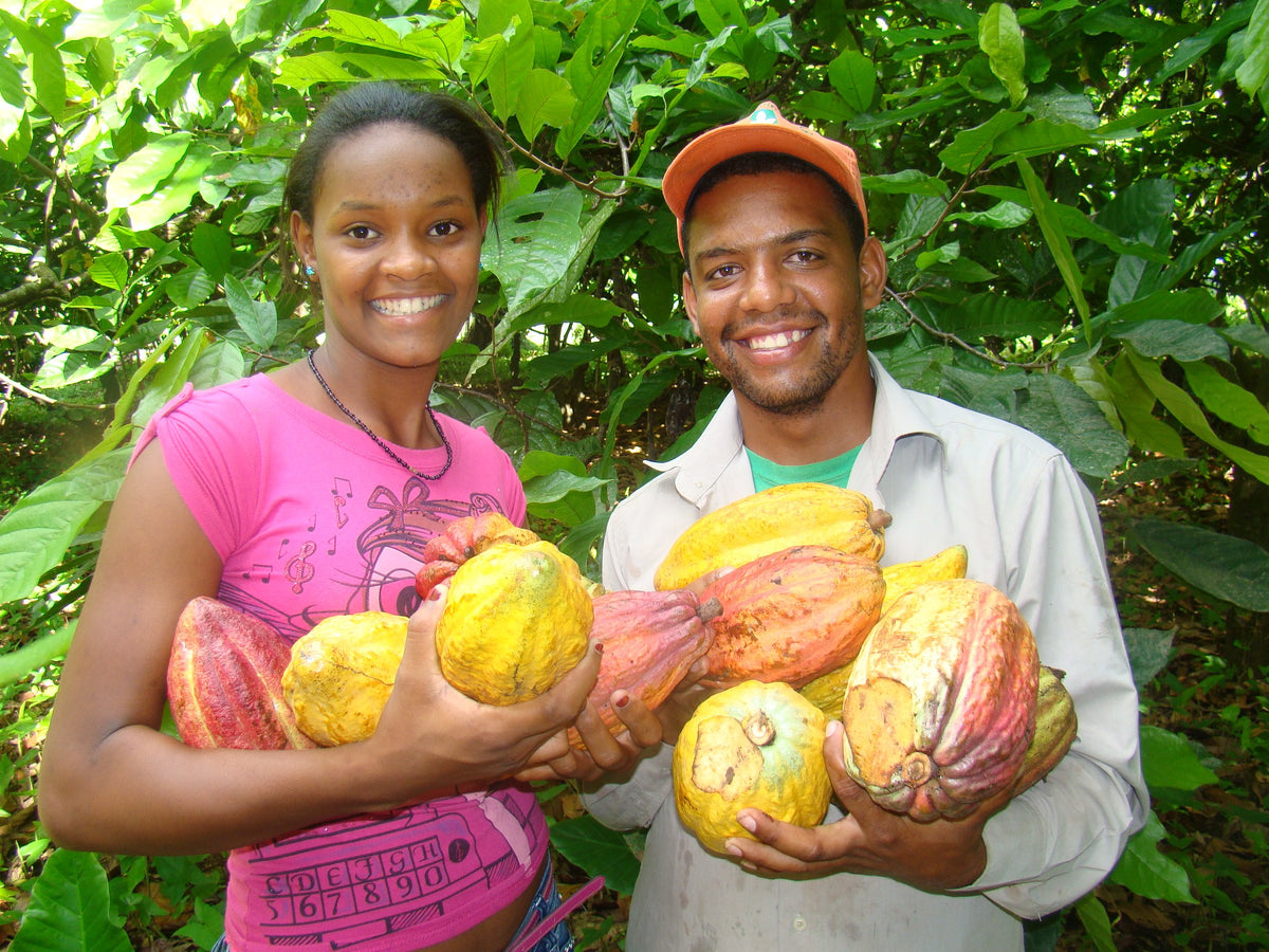 Una joven y un joven sonríen en un cacaotal, con los brazos llenos de mazorcas amarillas, naranjas y rojas